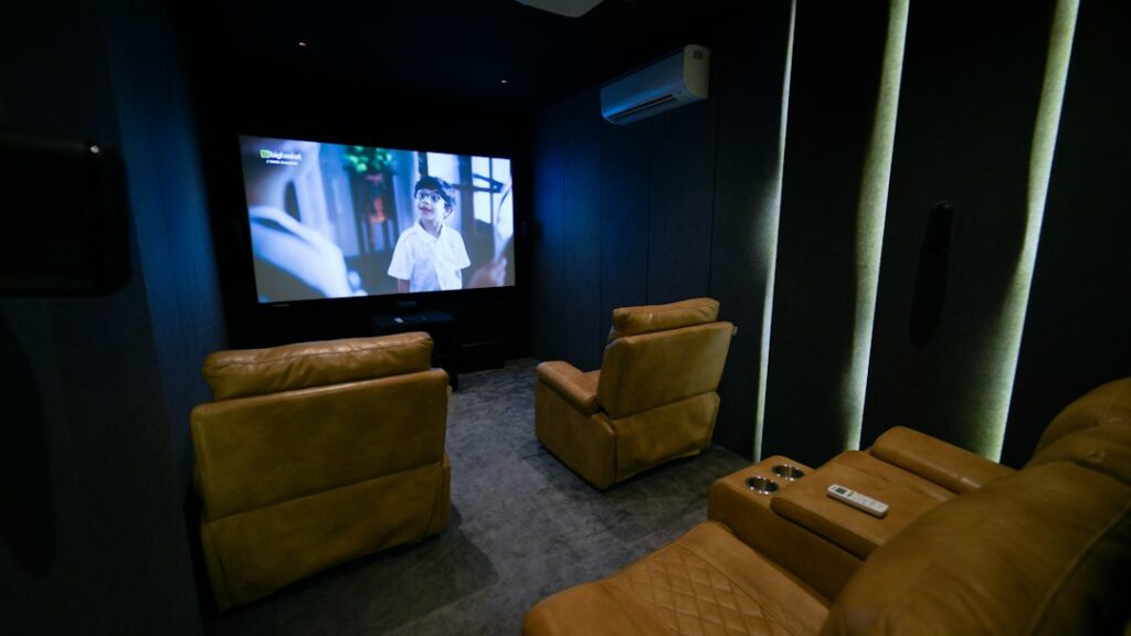 A finished theater room with tiered seating, a large screen, wall speakers, dim lighting, and acoustic panels.